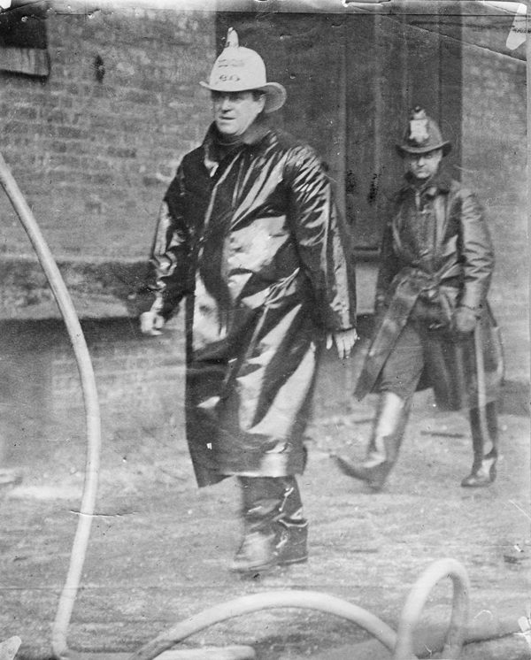 Fire Chief James J. Horan in white helmet and rubber coat walking past a building at a fire, Chicago Daily News, 1910.